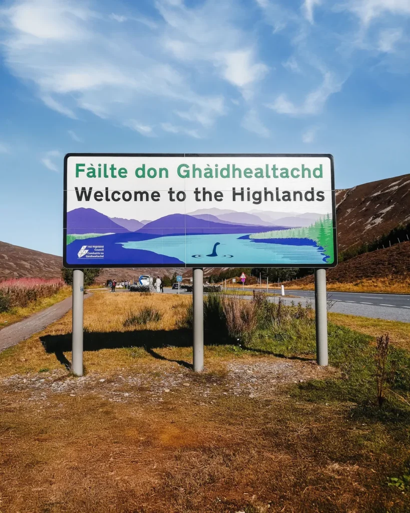 Straßenschild „Welcome to the Highlands" mit Loch-Ness-Illustration an der Grenze zum schottischen Hochland